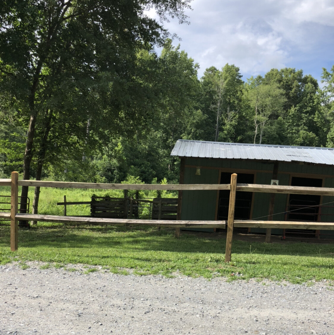 Basswood+Creek+Farms+NC+-+stable Rustic wooden fence in front of a green barn surrounded by lush trees and open grassland on a sunny day.